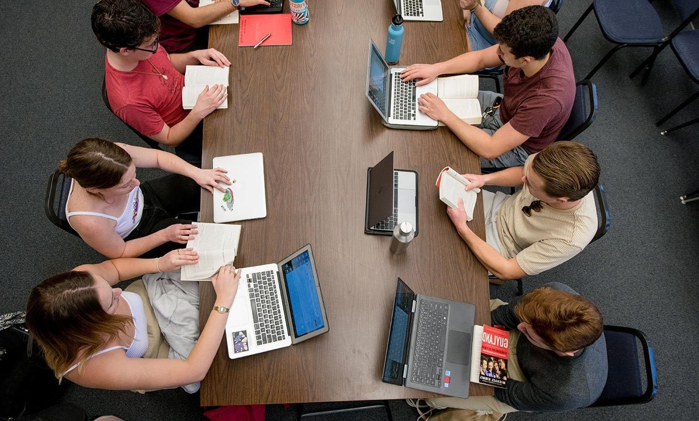 several students sit at a table with books and laptops open